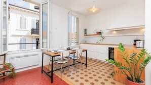 A compact kitchen with a dining area, large windows, and a potted plant. - Upstays République 67m² Apartment (Cannes)