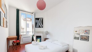 A hotel room with a large bed, a desk with a chair, a fireplace, and a view of the city through a window. - Upstays République 67m² Apartment (Cannes)