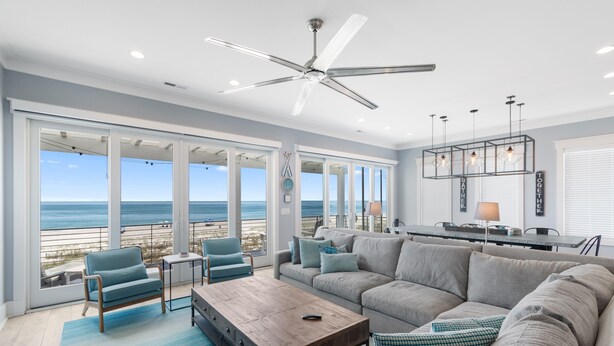 A spacious living room with a large window overlooking the beach, a ceiling fan, and a modern kitchen area.