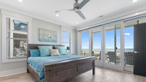 A bedroom with a large bed, a ceiling fan, and a view of the beach through the sliding glass doors.