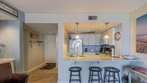 A modern kitchen with a central island, bar stools, and a view of the living area. - Ocean's Edge Retreat - St. Regis Condo #1109 (North Topsail Beach)