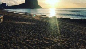 Een strand met een opvallende rotsformatie in de achtergrond en de zon die ondergaat boven de oceaan.