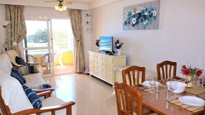 Un salon moderne avec un canapé, des fauteuils, une table à manger pour quatre personnes, un téléviseur posé sur un meuble et un balcon avec des rideaux.