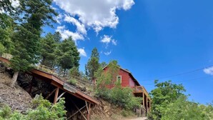 A red wooden house with a dirt path leading to it, surrounded by greenery and a clear blue sky. - Secluded Mountain Cabin: Hot Tub & Spectacular (Ruidoso)