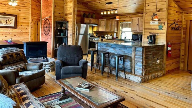 A cozy log cabin interior with a living room, a fireplace, a kitchen with a bar, and a stone fireplace. - Secluded Mountain Cabin: Hot Tub & Spectacular (Ruidoso)