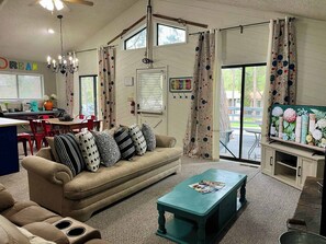 A cozy living room with a sofa, coffee table, and a television. There is a dining area with chairs and a table, and a kitchen area with a coffee maker and a vase.