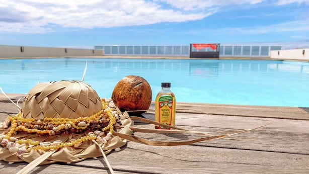 En træterrasse med en flettet kurv, en kokosnød og en flaske Monoi Tiki Tahiti. En swimmingpool ses i baggrunden.