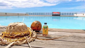 A wooden deck with a woven basket, a coconut, and a bottle of Monoi Tiki Tahiti. A swimming pool is visible in the background.