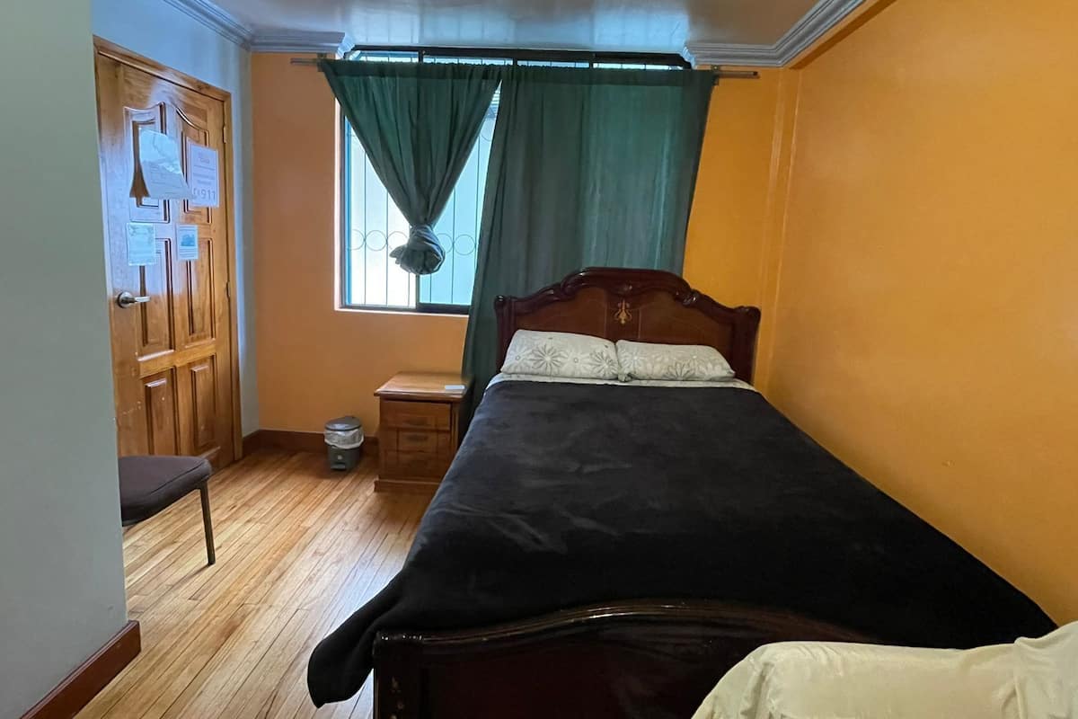 A small, narrow room with a bed, a wooden chair, a small table, a window with curtains, and a door with a sign.