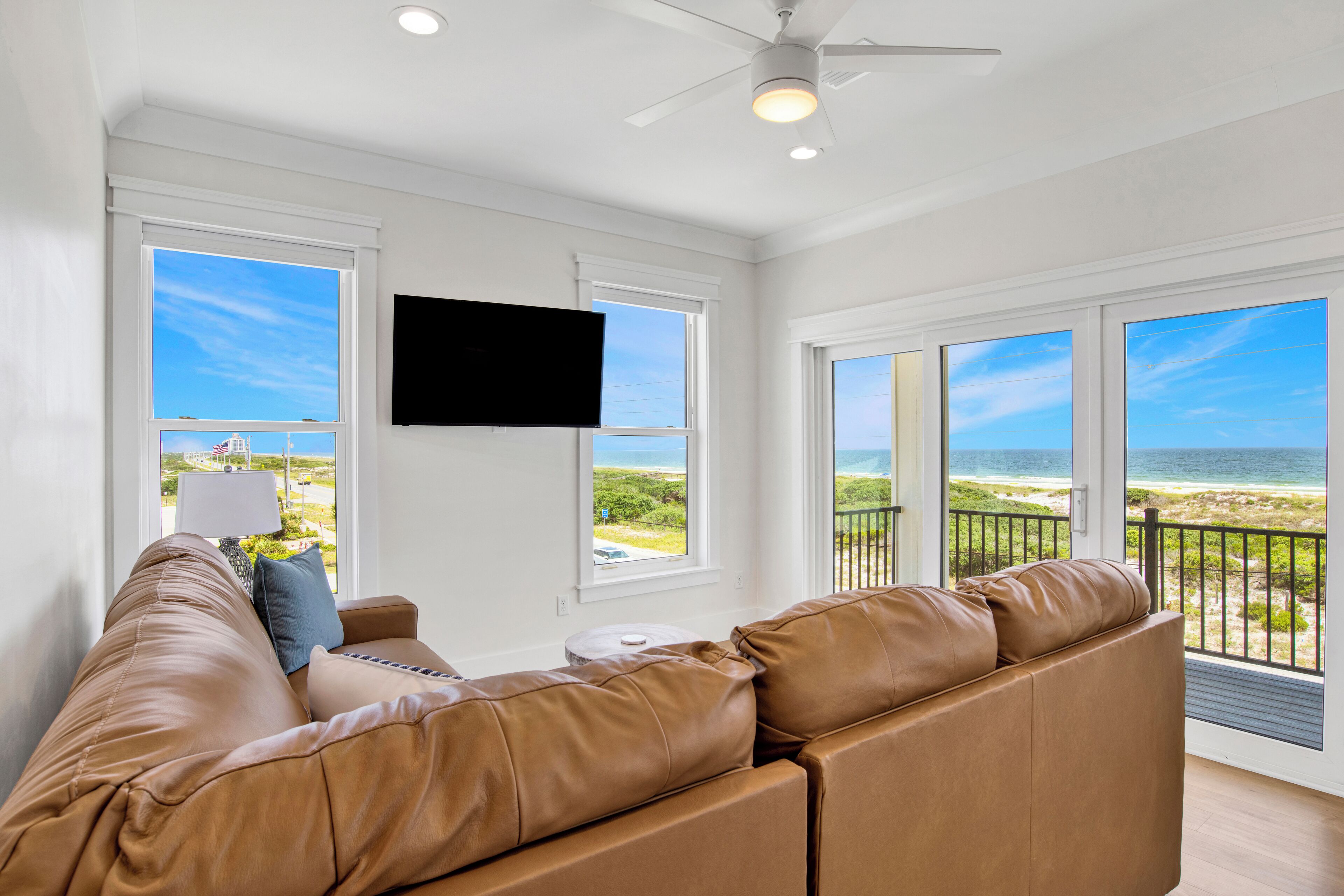 A spacious living room with a large leather sofa, a flat-screen TV mounted on the wall, and large windows offering a view of the beach.