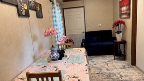 Een eetgedeelte met een tafel bedekt met een bloemrijk tafelkleed, een vaas met roze bloemen en een blauw-wit keramieken krukje. Op de achtergrond staat een donkerblauwe bank en een klein houten tafeltje met een vaas rode bloemen.