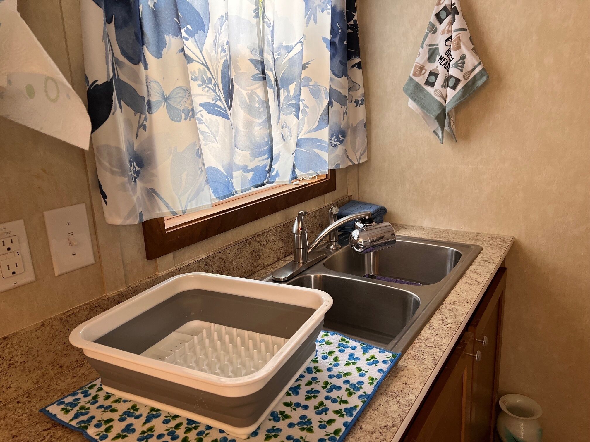 A kitchen with a double sink, a drying rack, and a window with a floral curtain.