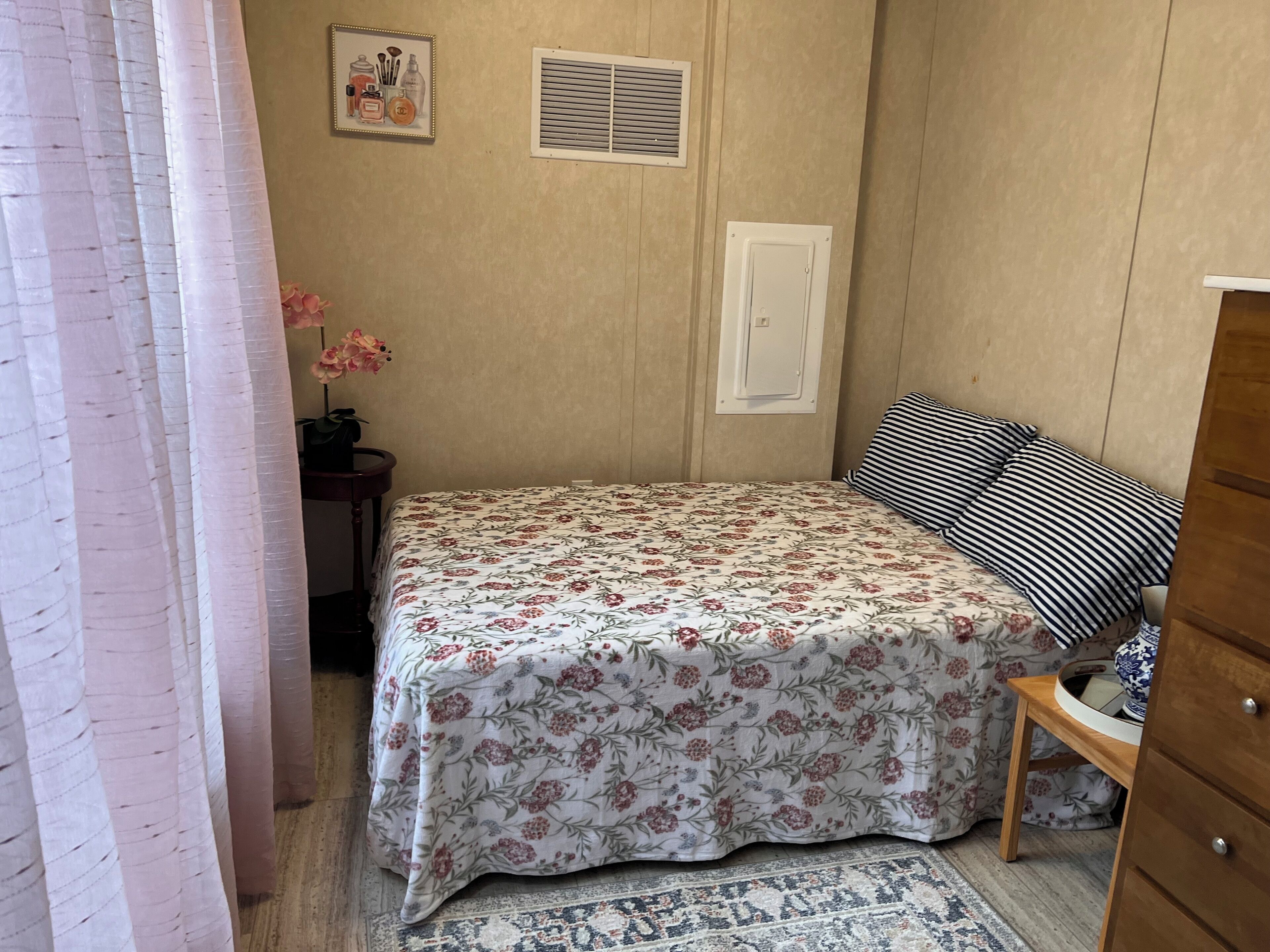 Une chambre avec une couvre-lit à motifs floraux, une commode en bois, un coussin rayé, une petite table avec un vase et une grille de ventilation fixée au mur.