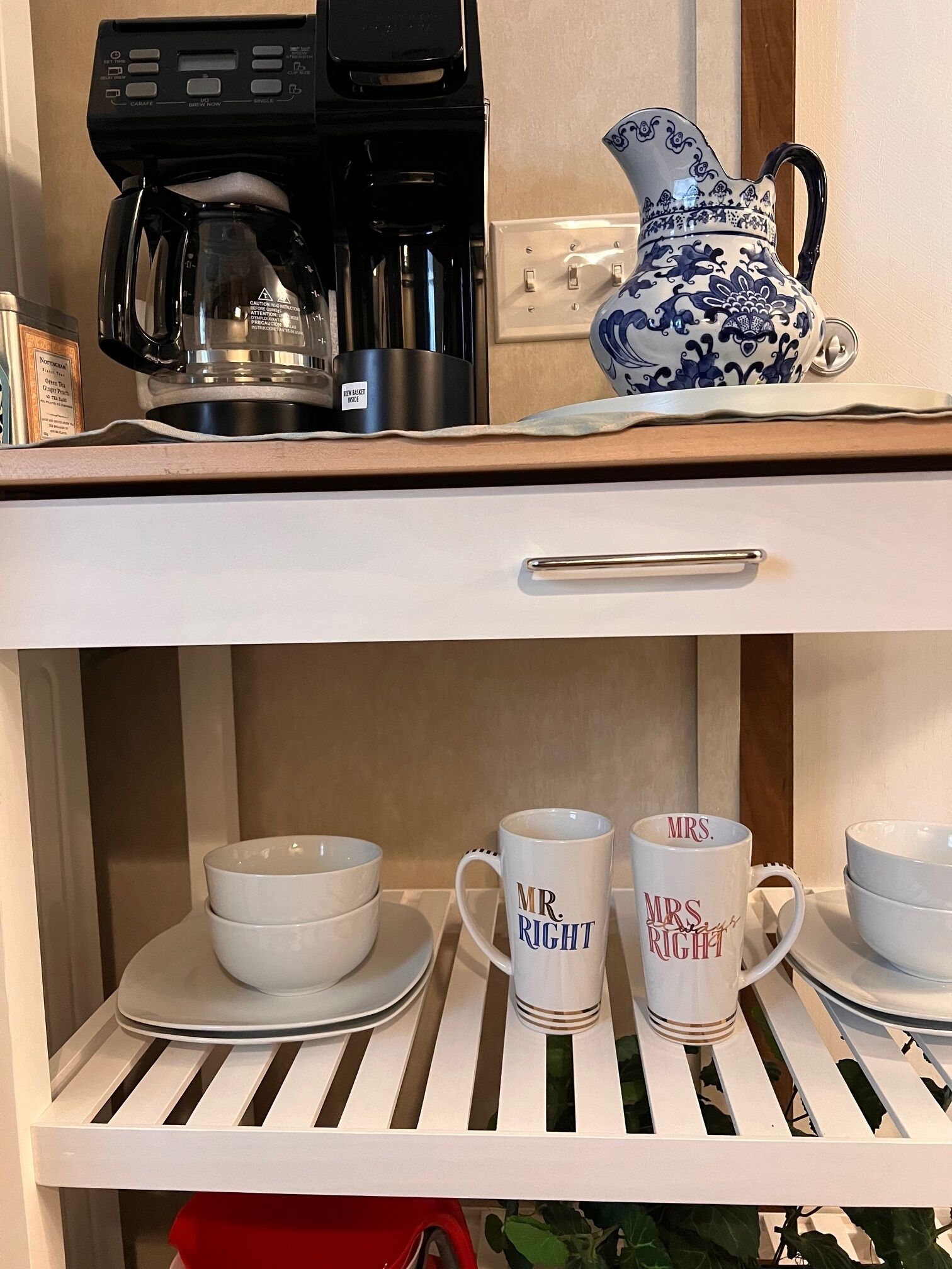 Un taulell de cuina amb una cafetera, una gerra blava i blanca, i un safata blanca. A sota, hi ha dos vasos blancs amb la inscripció "Mr. Right" i "Mrs. Right" sobre una estant blanca.