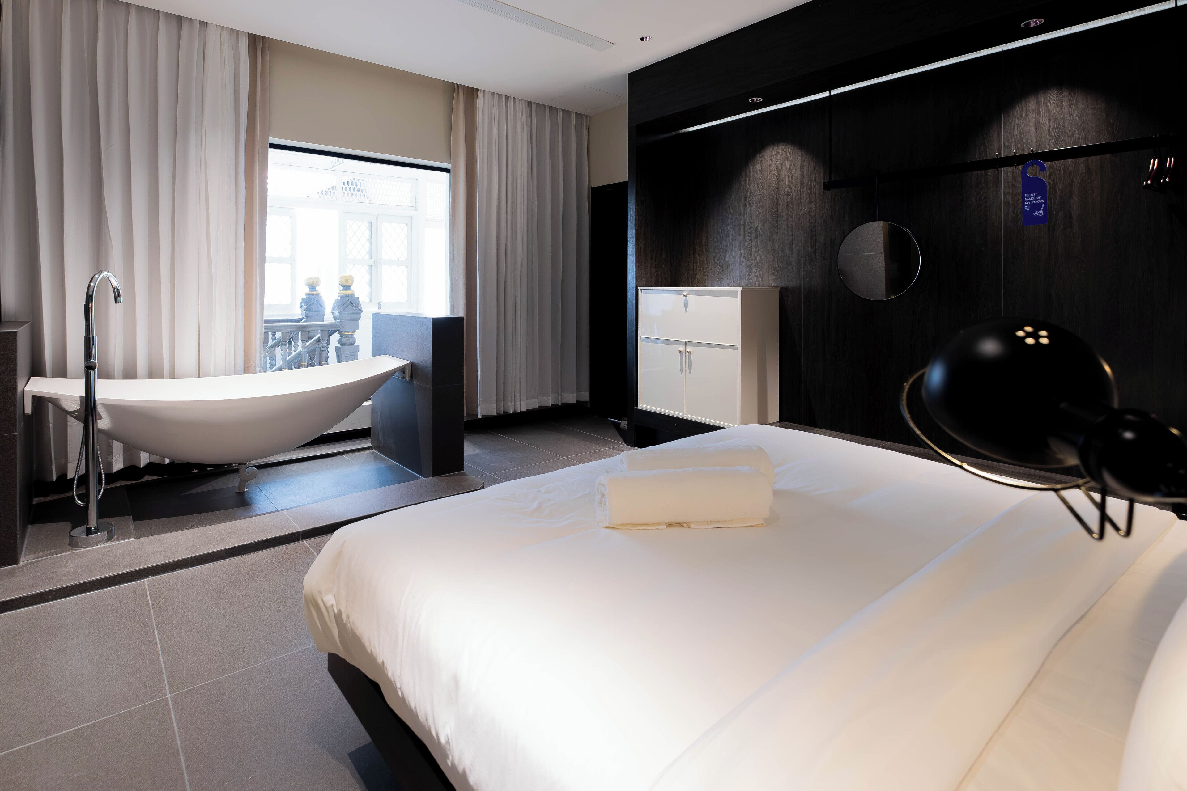 A modern hotel room with a large bed, a bathtub, and a view of a balcony with two people.