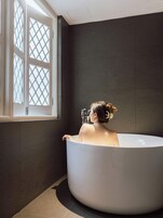 A woman in a bathtub in a room with dark walls and a window with a lattice design.