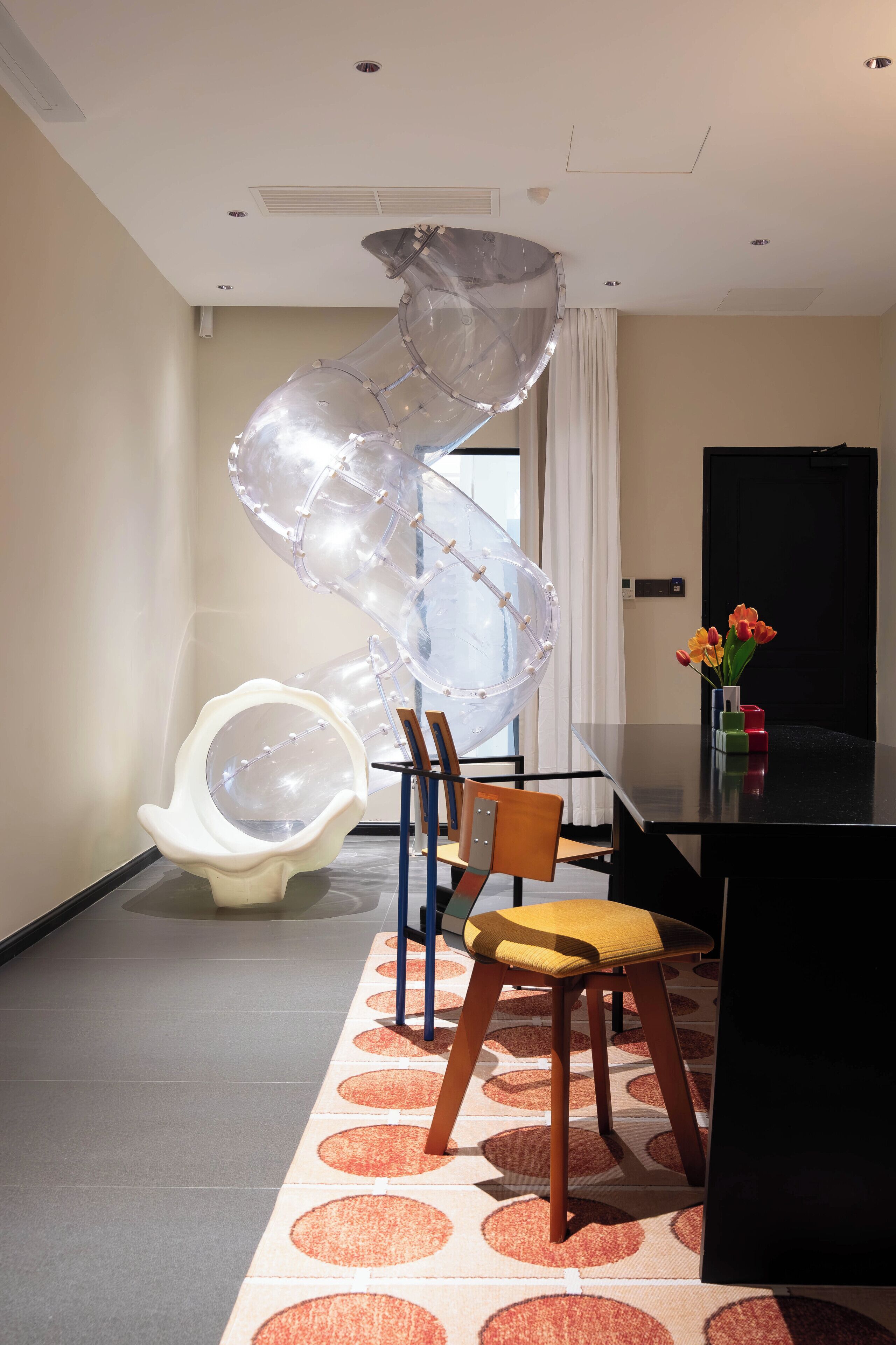 Een moderne hotelkamer met een spiraalvormige glaskunst, een witte stoel, een houten stoel, een zwarte tafel en een vaas met bloemen.