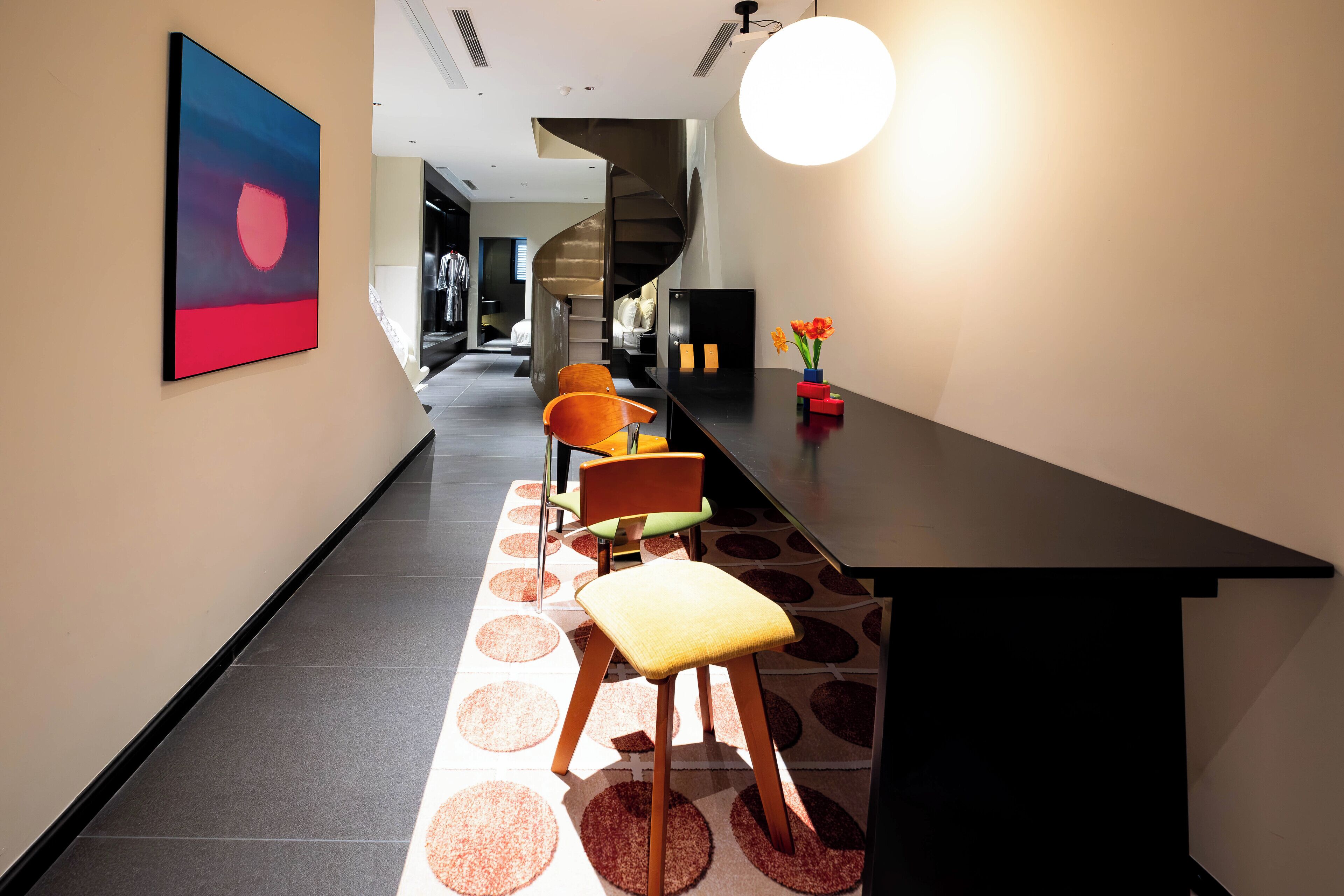 A modern interior with a long hallway, a spiral staircase, and a dining area with a black table and colorful chairs.
