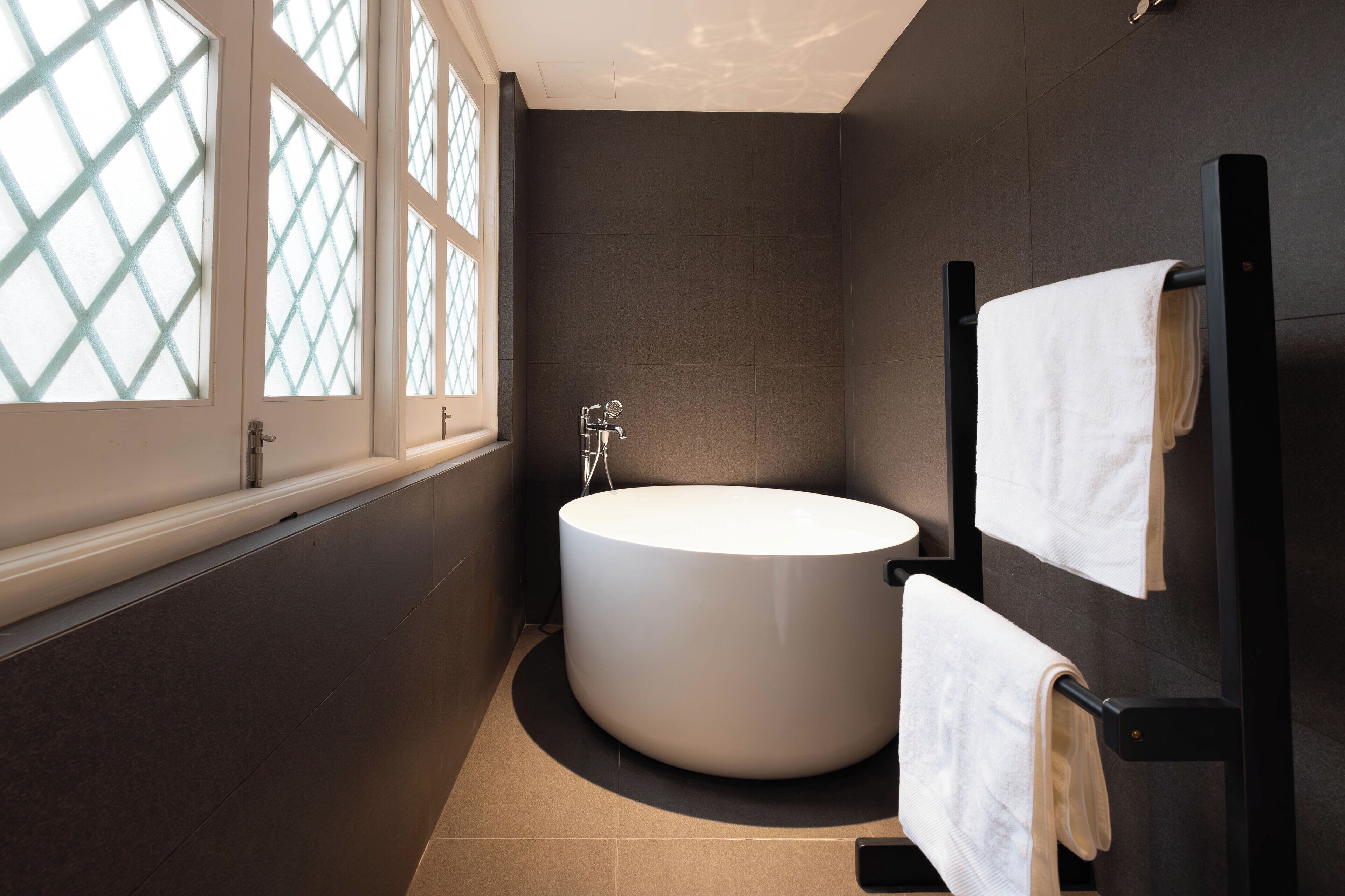 A modern bathroom with a freestanding bathtub, a towel rack, and a window with a diamond-patterned design.