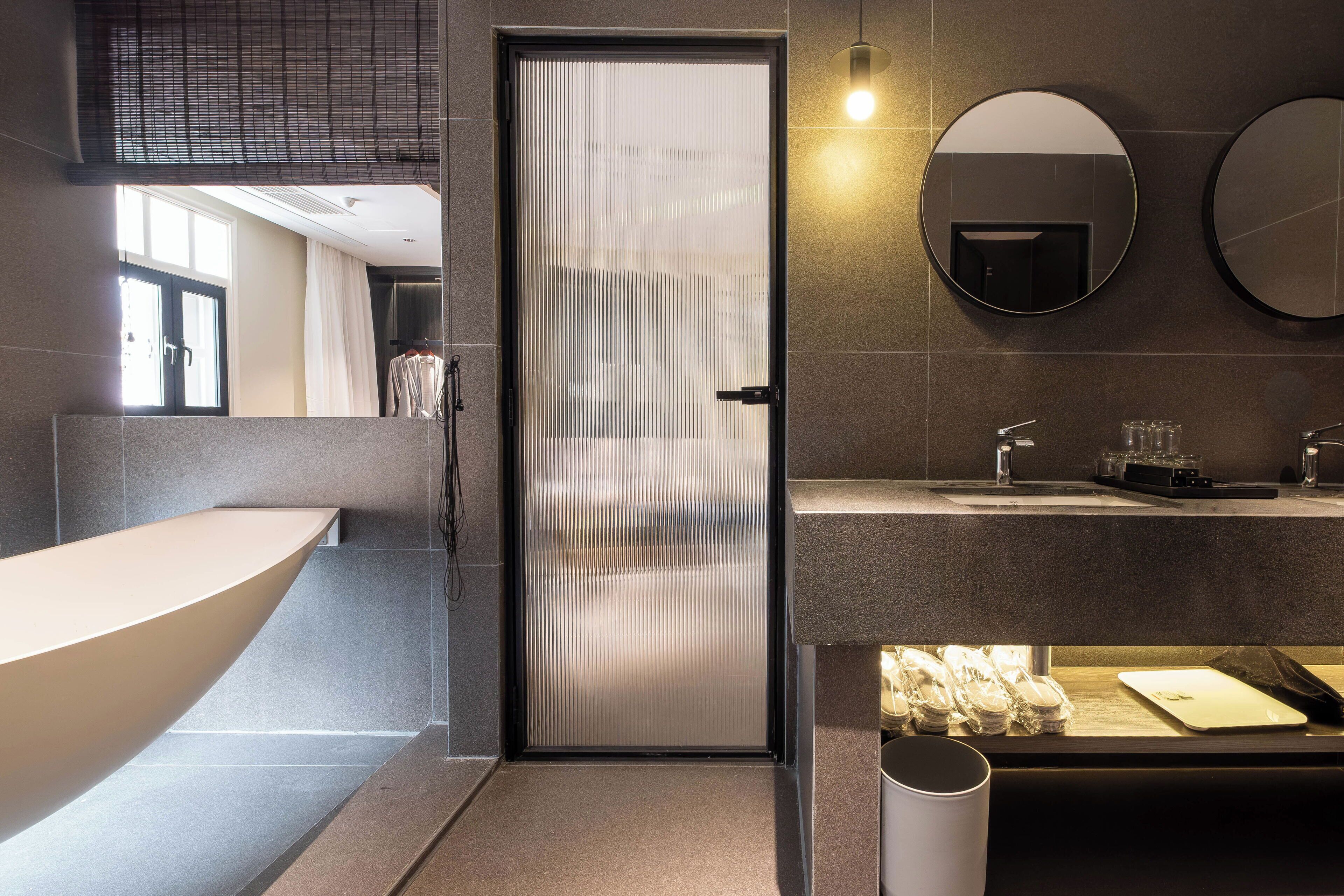 Ein modernes Badezimmer mit einer großen Badewanne, einer Toilette und einem Waschbecken. Zwei Spiegel sind an der Wand angebracht, und ein Fenster sorgt für natürliches Licht.