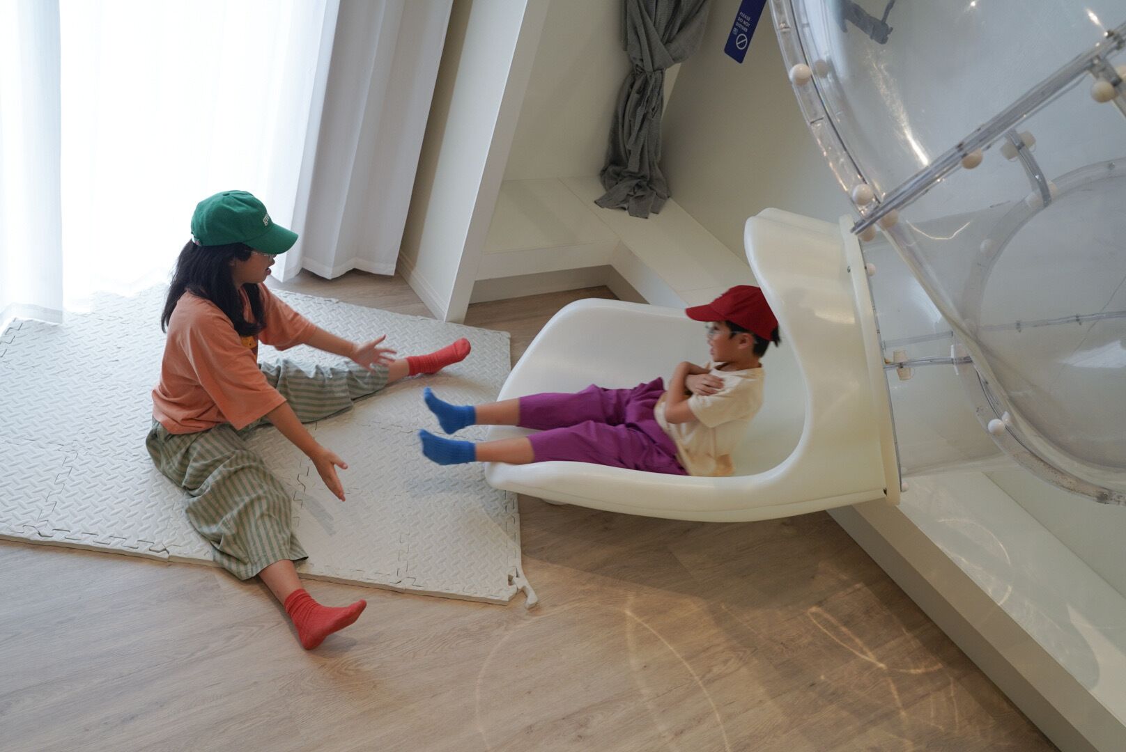 Dos niños jugando en una habitación con una bañera blanca, piso de madera y una cortina blanca.