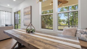 A modern living room with a wooden dining table, large windows, a ceiling fan, and a view of trees. - Seaside Serenity Boutique Home (Gulfport)