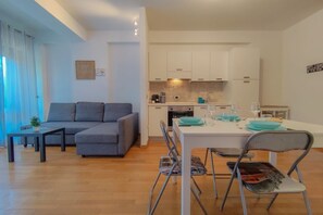Un salon moderne avec un canapé gris, une table à manger avec des chaises et un coin cuisine doté de meubles blancs.