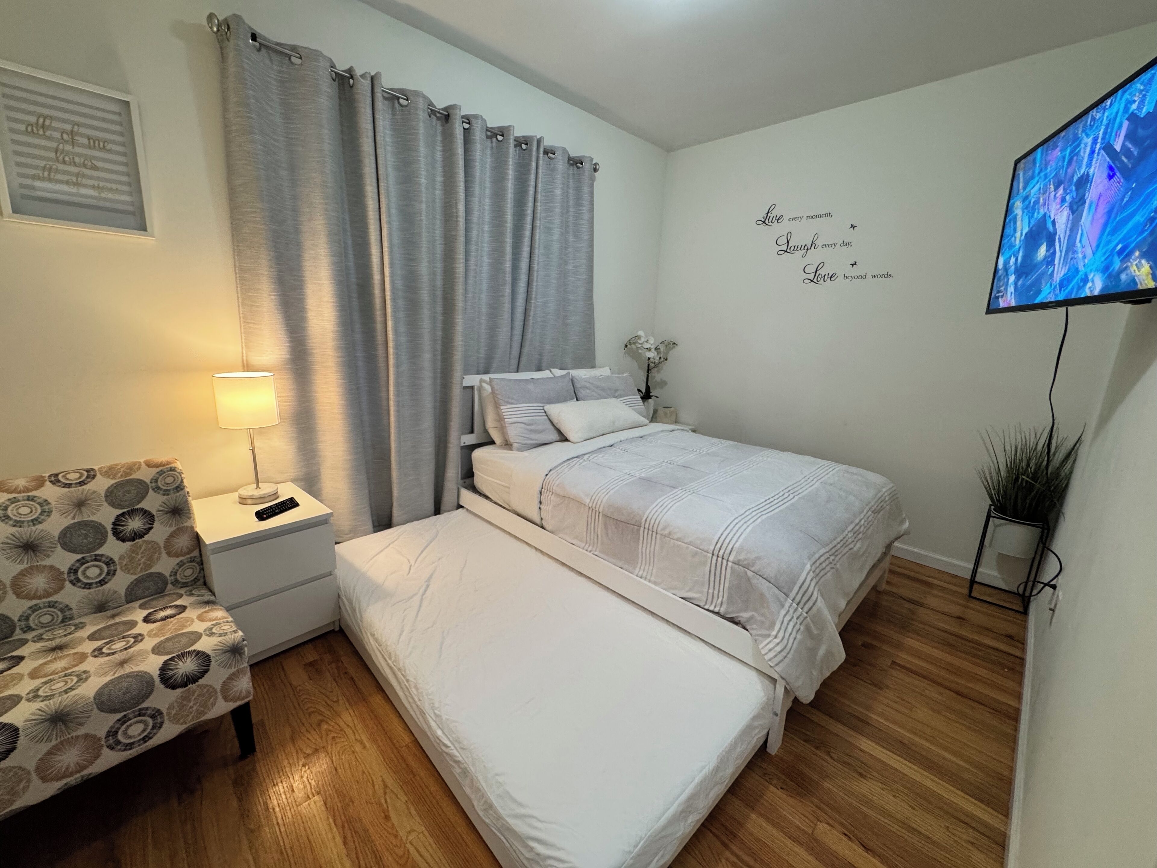 Un dormitorio con cama, una silla, mesita de noche con lámpara, televisor y una pared con citas motivacionales.