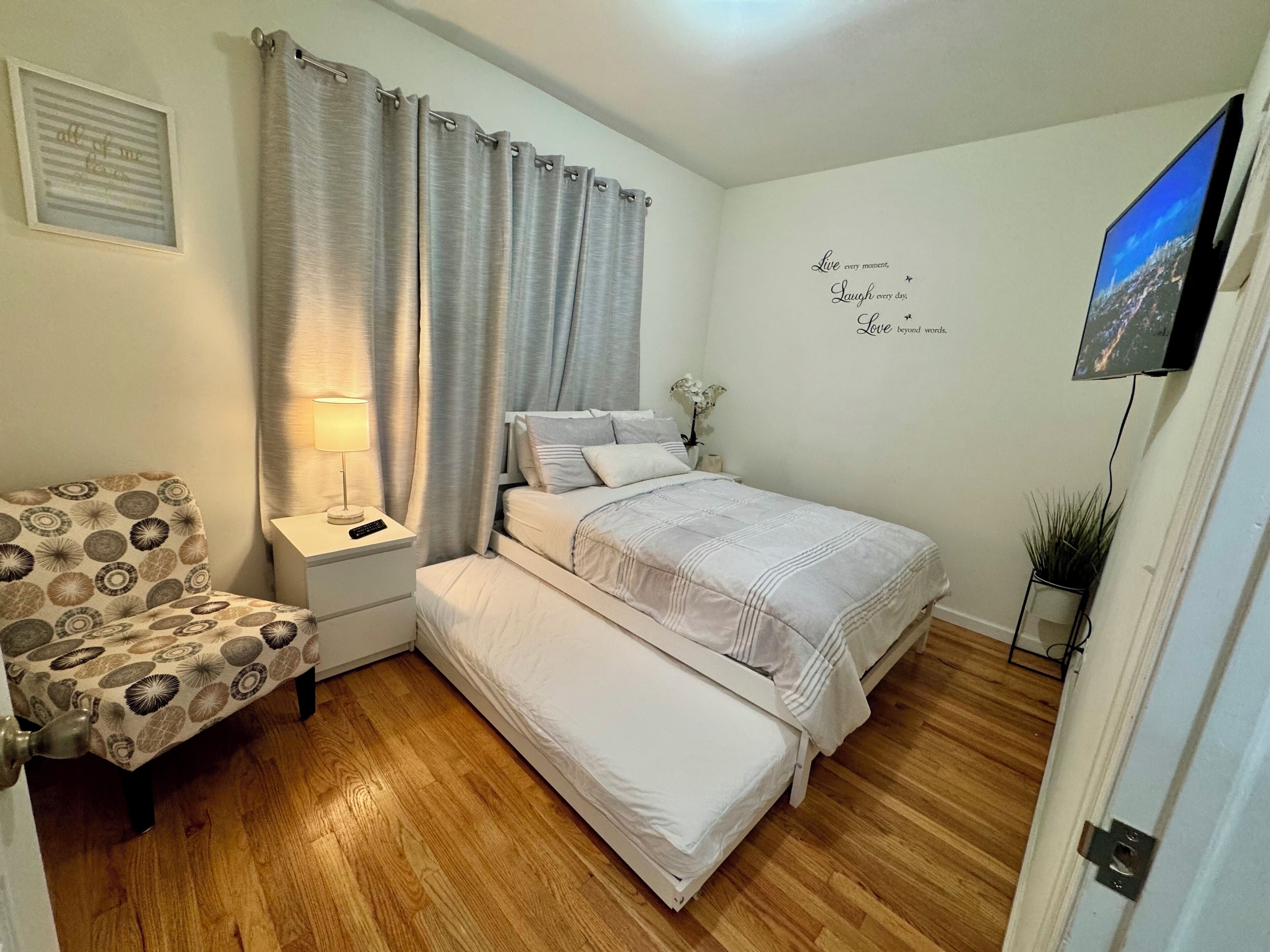 Quarto com cama, mesa de cabeceira, abajur, cadeira e televisão na parede.