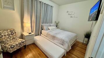 Quarto com cama, mesa de cabeceira, abajur, cadeira e televisão na parede.