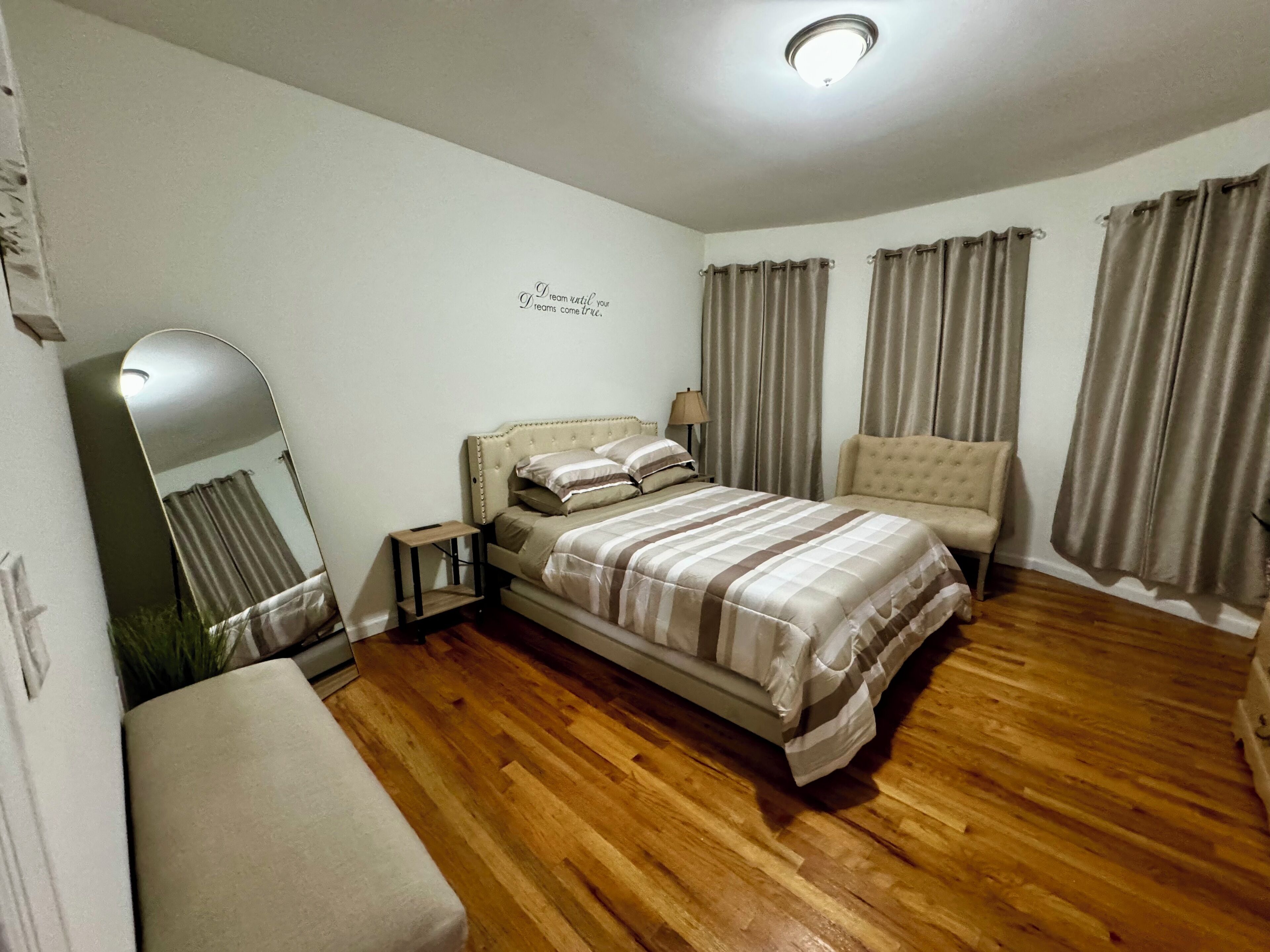 Een slaapkamer met een bed, een stoel, een tafeltje, een spiegel en gordijnen.