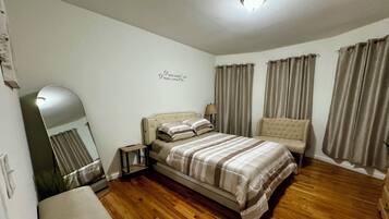 Een slaapkamer met een bed, een stoel, een tafeltje, een spiegel en gordijnen.