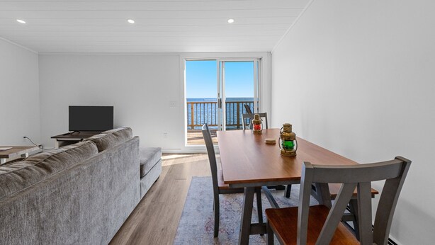 Un salon moderne comprenant un canapé, un téléviseur, une table à manger avec des chaises, et une vue sur la mer grâce à une grande fenêtre.