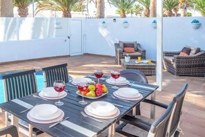 An outdoor patio with a dining table set for four, surrounded by wicker furniture, a pool, and palm trees. - Villa Canada (Tías)
