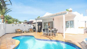 A swimming pool with a tiled edge, a covered patio area with a table and chairs, and a white building with a covered entrance. - Villa Canada (Tías)