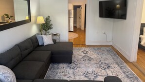 A modern living room with a sectional sofa, a large flat-screen TV, a ceiling fan, and a patterned rug.