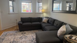 A living room with a sectional sofa, a patterned rug, a round coffee table with remotes, and a large mirror on the wall.