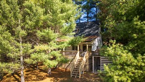 Une cabane entourée d’une dense forêt de pins, dotée d’une terrasse en bois et d’un escalier menant à l’entrée.