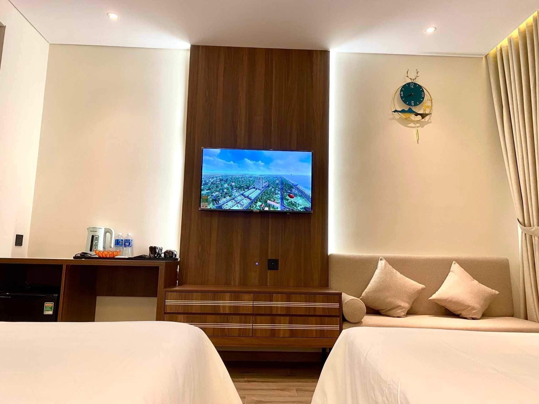 A hotel room with a TV mounted on a wooden panel, a sofa with cushions, and a desk with a kettle and water bottles.