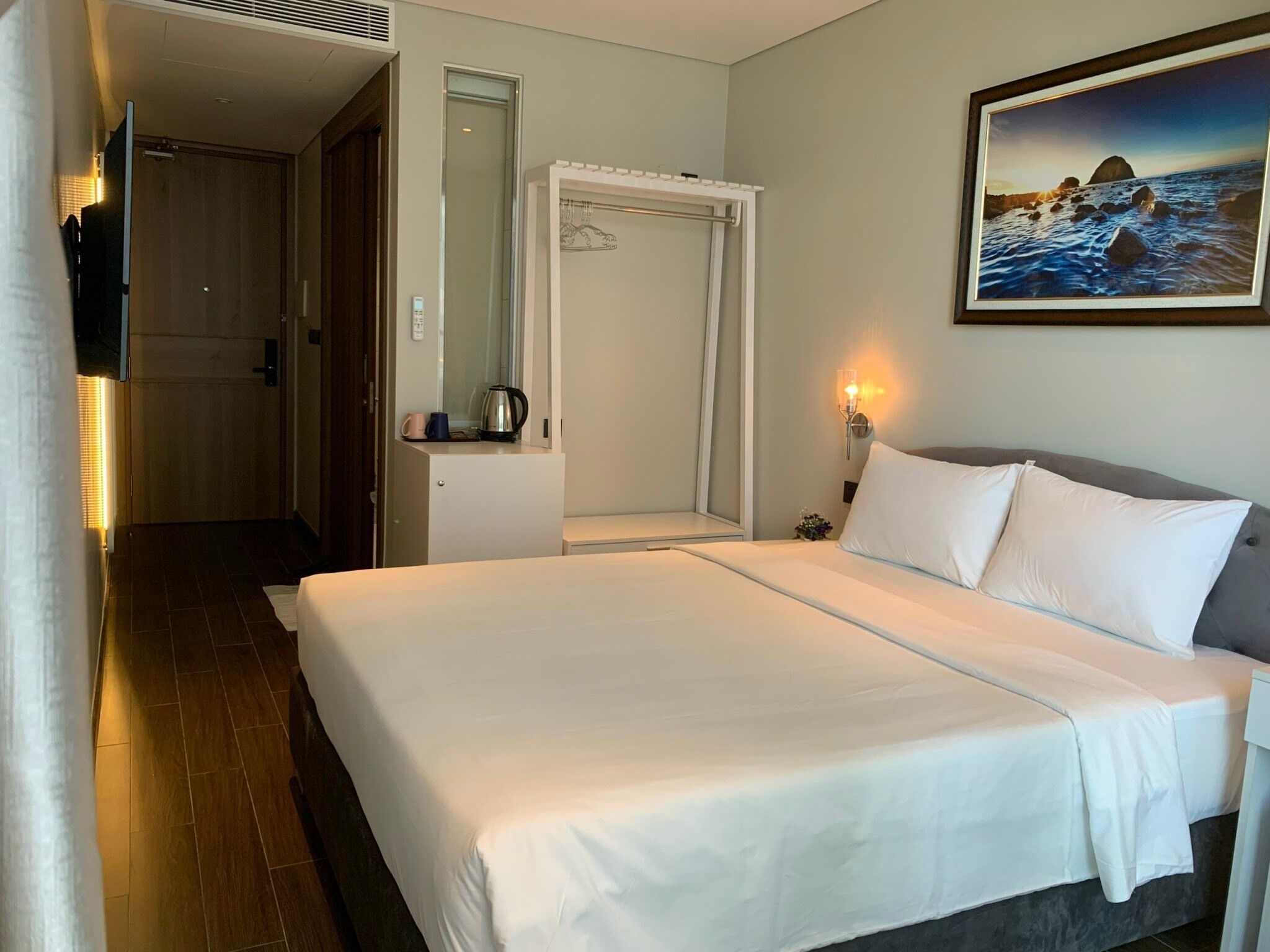 A hotel room with a bed, a nightstand, a wall-mounted TV, a painting, and a door leading to another room.