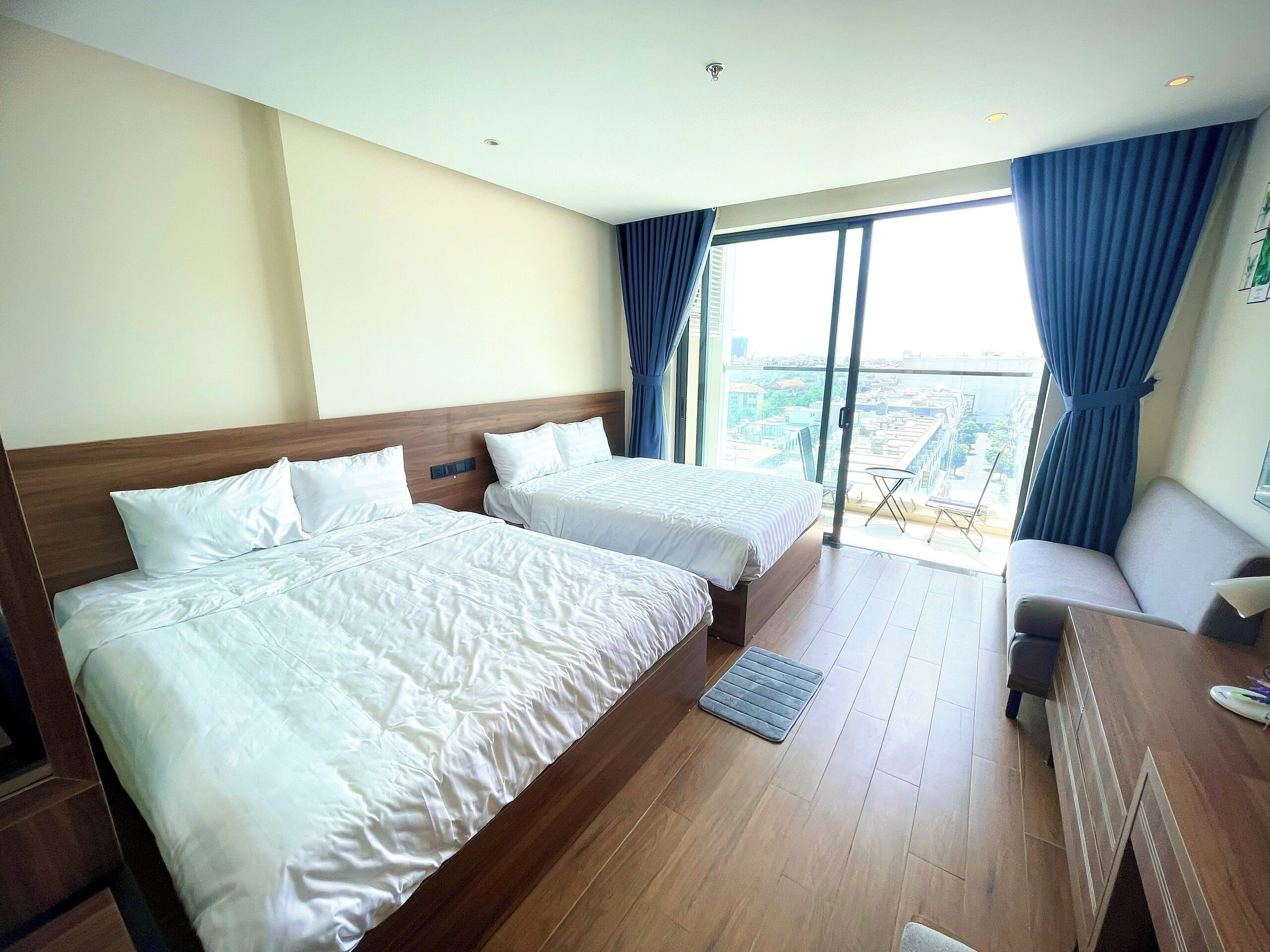 A hotel room with two beds, a sofa, a small table, and a view of the city through large windows.