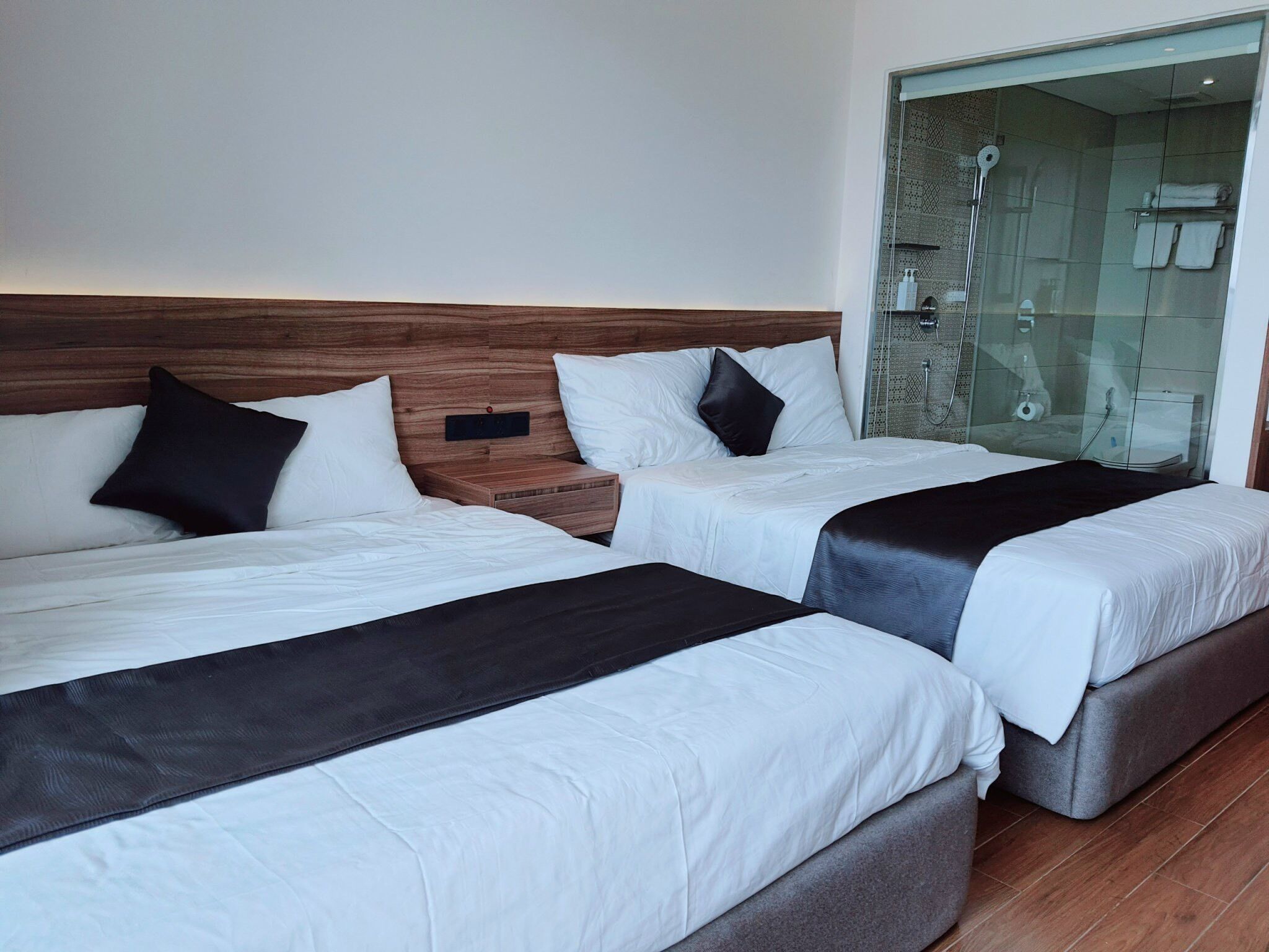 A hotel room with two beds, a wooden headboard, a glass shower enclosure, and a small bedside table.
