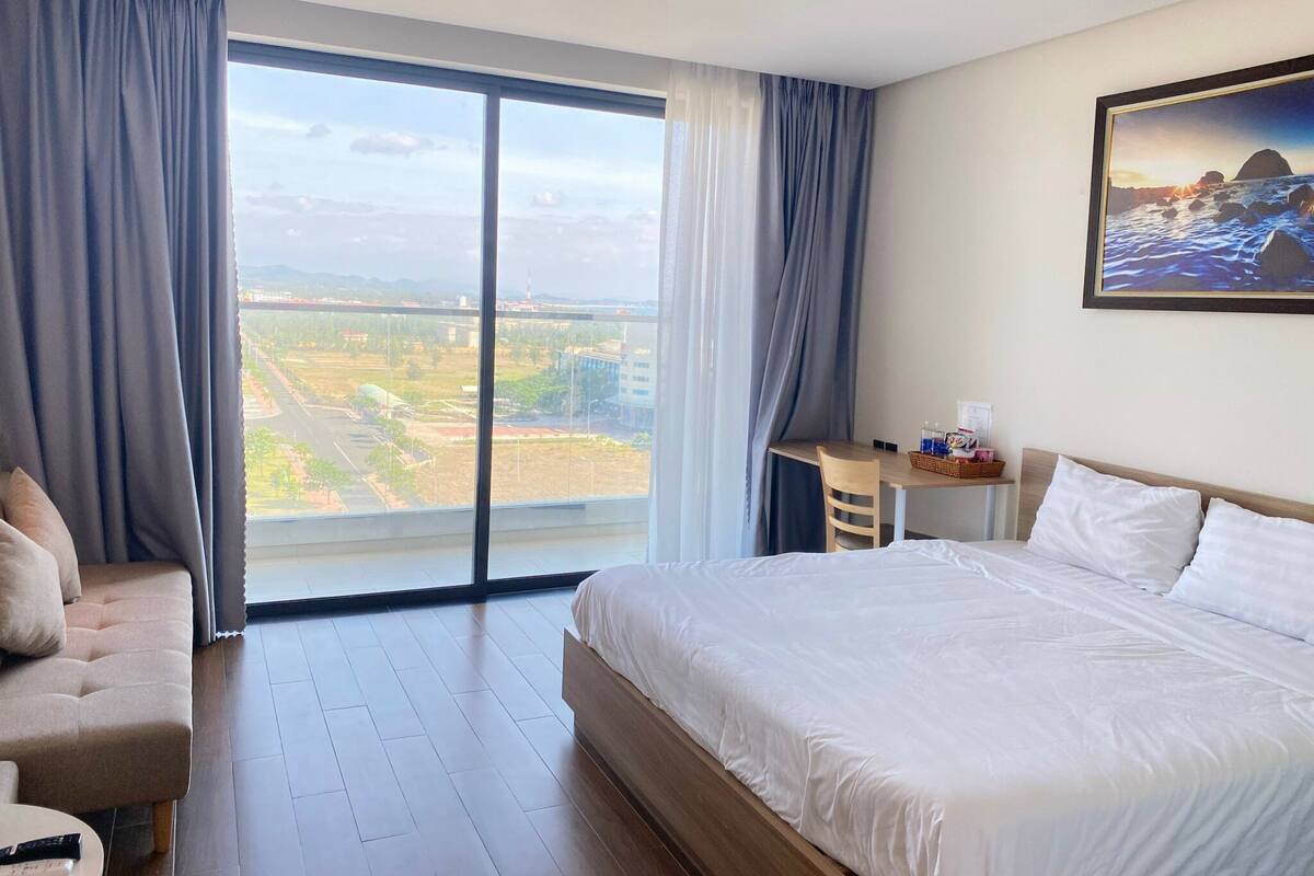 Habitación de hotel con una cama grande, un escritorio y vistas a un paisaje urbano a través de ventanas de piso a techo.