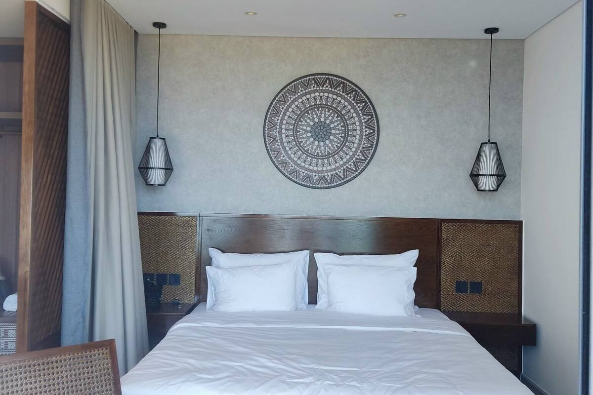 Una habitación de hotel moderna con una cama grande, dos mesitas de noche con lámparas, un tapiz decorativo en la pared y vistas al exterior.
