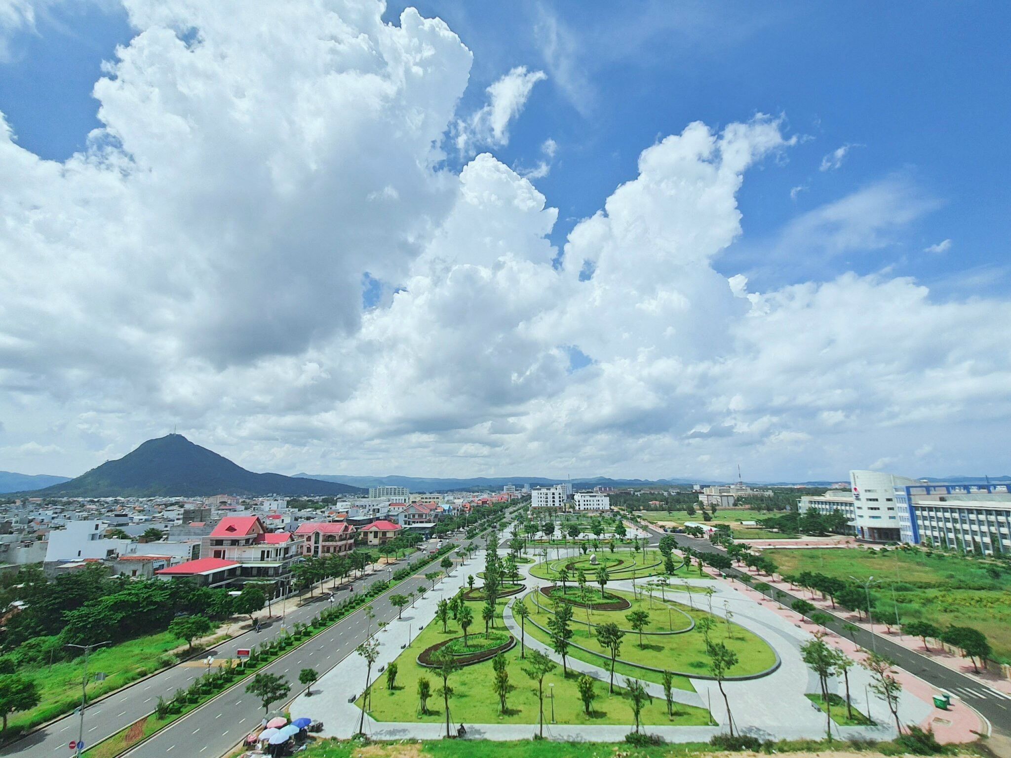 A cityscape with a prominent mountain in the background, a wide road with greenery and trees, and a clear sky with clouds.