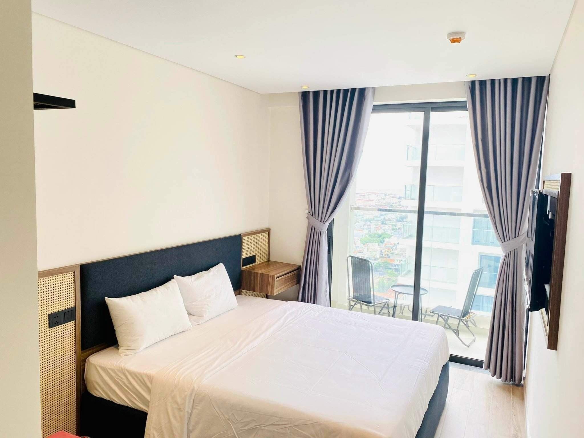 Quarto de hotel com uma cama grande, uma mesinha, uma cadeira e uma varanda com vista para a cidade.