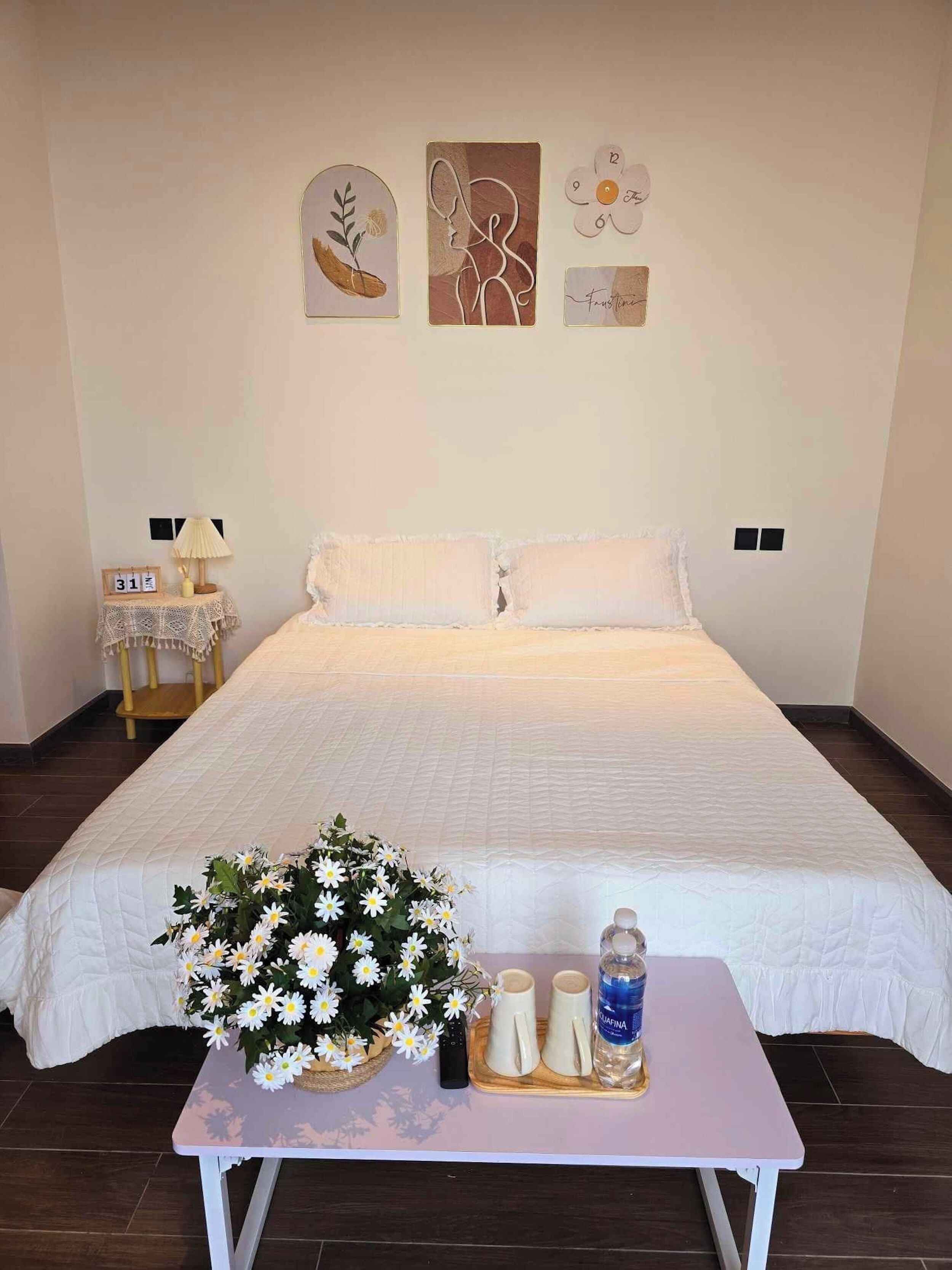 A bedroom with a large bed, a bedside table with a vase of flowers, and a small table with two cups and a bottle.
