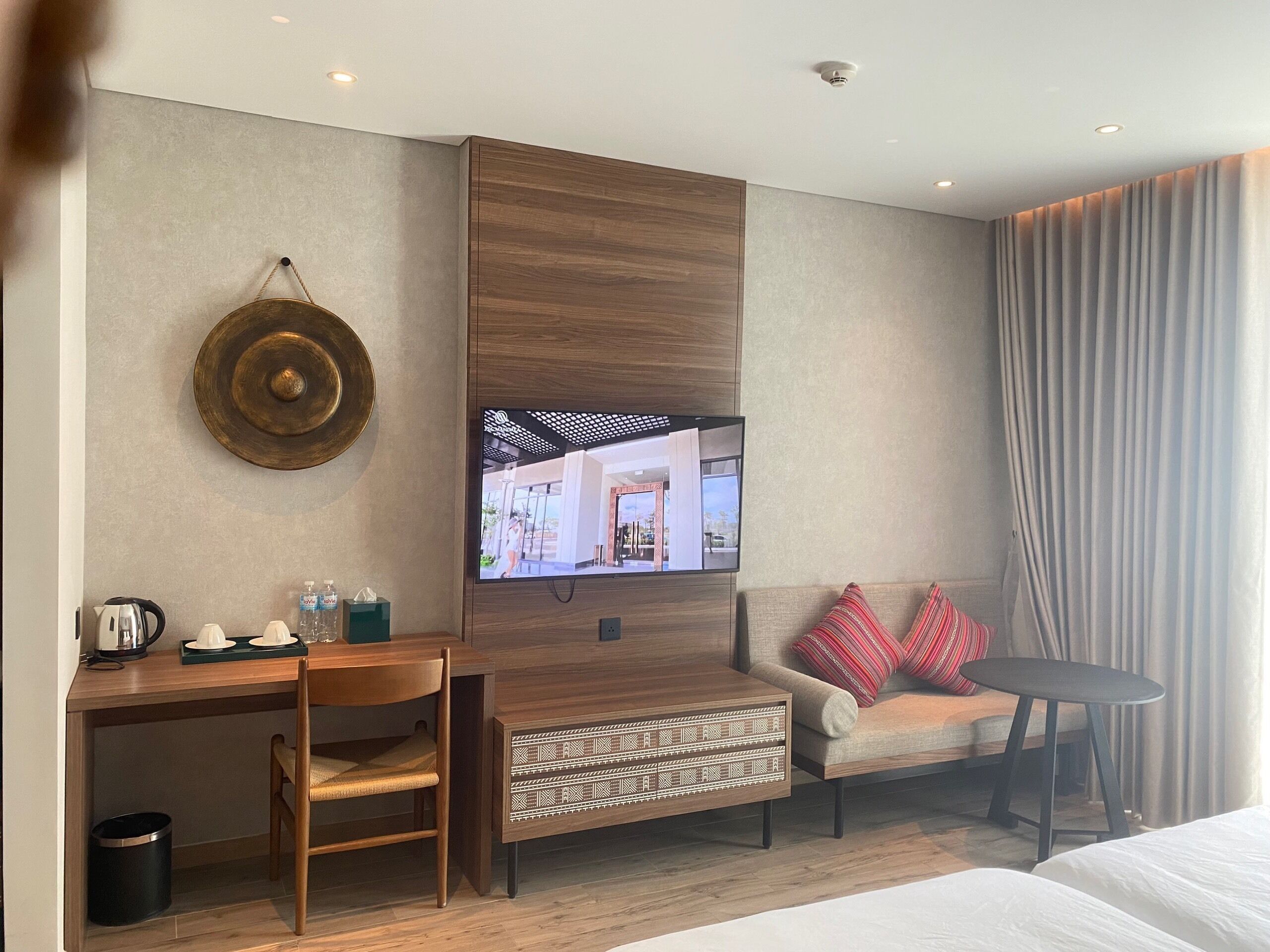 Hotelkamer met een houten bureau, een stoel, een bank met rode kussens, een aan de muur bevestigde televisie en een groot raam met gordijnen.