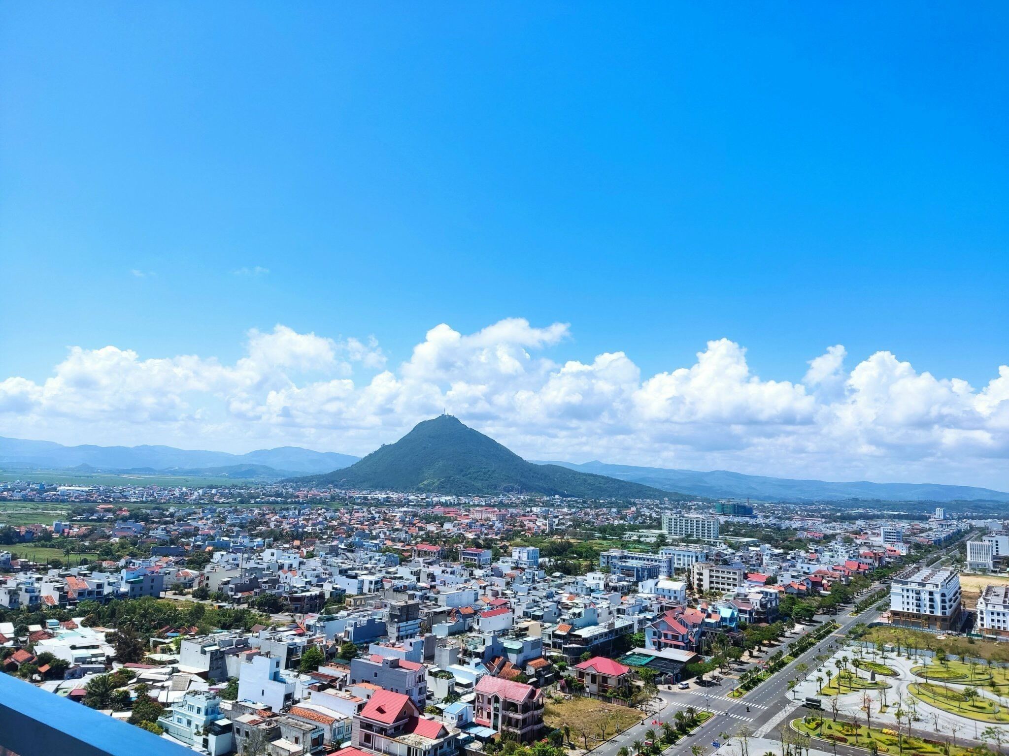 Un paisaje urbano con una montaña destacada al fondo, una zona densamente poblada con diversos estilos arquitectónicos y un cielo azul despejado.