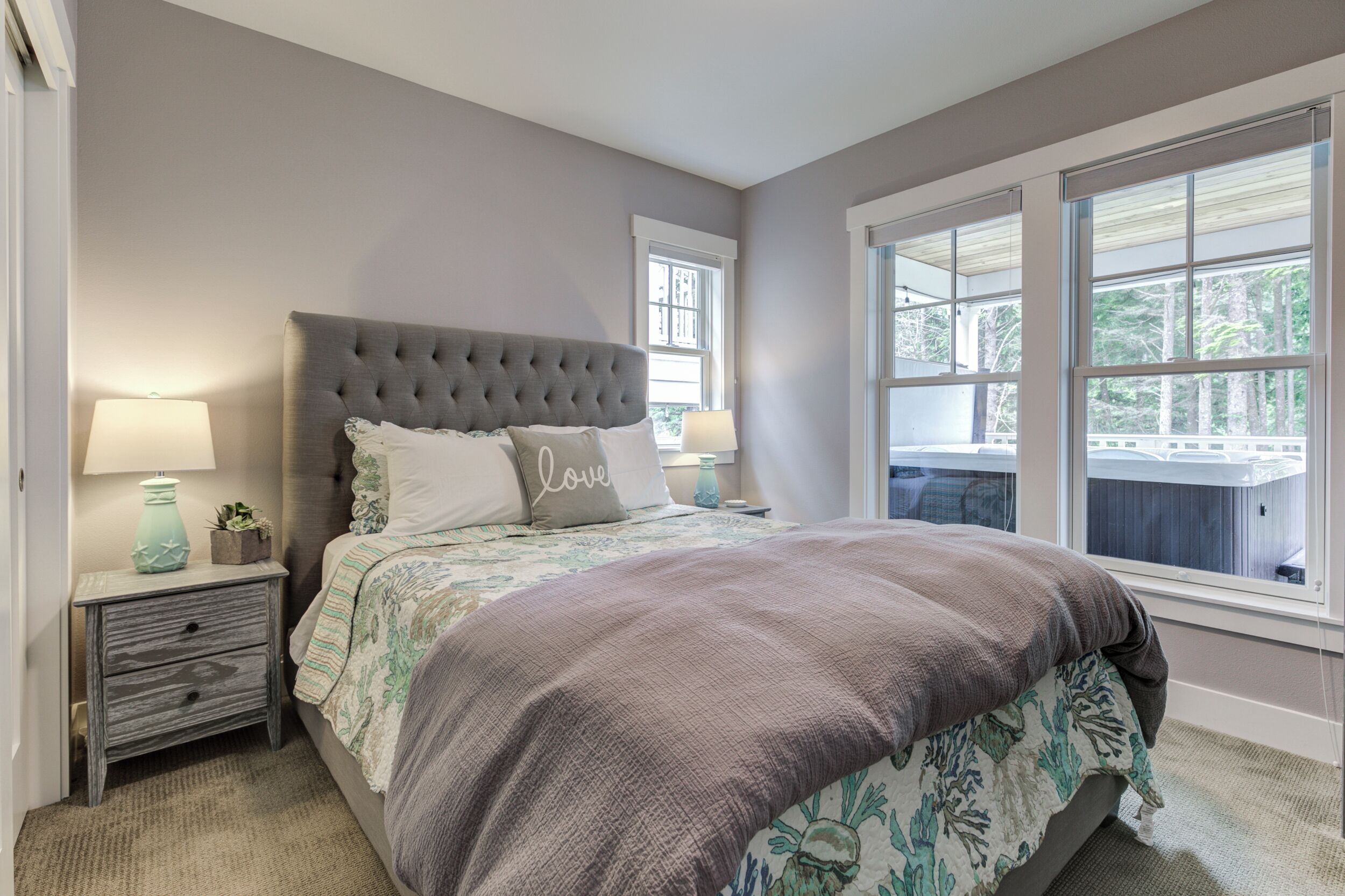 A bedroom with a tufted headboard, a bed with floral bedding, a nightstand with a lamp, and large windows offering a view of the outdoors.